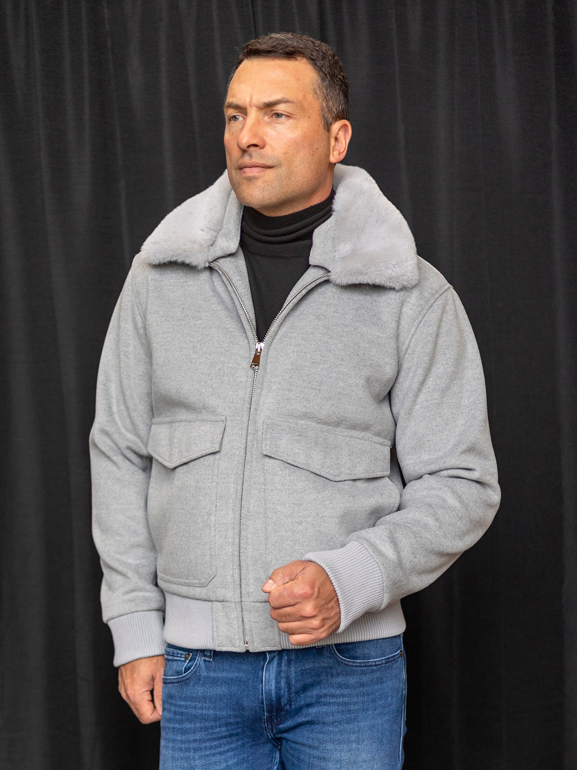 A man wearing the INSOMNIA MZW-501 Tailored Fit Grey Wool Bomber Jacket with a removable faux fur collar, paired with blue jeans, stands confidently in front of a black curtain.