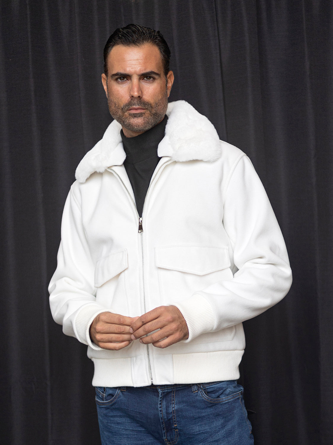 A person wearing the Insomnia MZW-501 Tailored Fit Wool Bomber Jacket in white, featuring a faux fur collar, stands in front of a dark curtain.