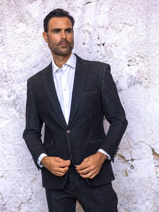 A man in the INSOMNIA NOBU 2PC Tailored Fit Black Denim Suit by Insomnia, paired with a crisp white shirt, stands against a textured, cracked wall, embodying timeless fashion.