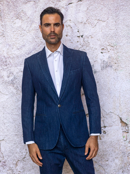 A man showcases his fashion sense in the INSOMNIA NOBU 2PC Tailored Fit Blue Denim Suit by Insomnia, paired with a crisp white shirt, as he stands against a textured, light-colored wall.