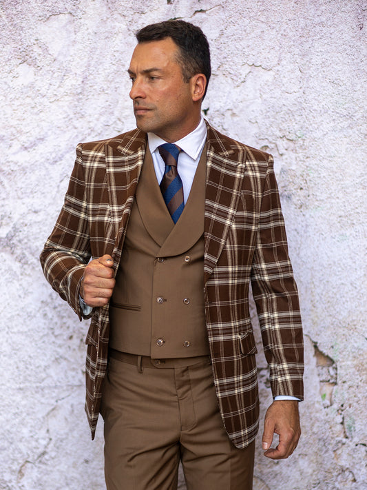 A man in an Insomnia tailored fit brown plaid jacket from the INSOMNIA PARIS-1 3PC Compose Tailored Fit Brown Suit, paired with a tan double-breasted vest and pants, accessorized with a blue and brown striped tie, stands against a textured light-colored wall, perfectly composing an impeccable ensemble.