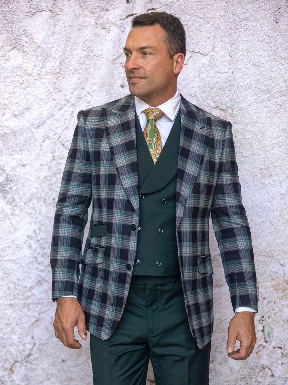 A man showcases the INSOMNIA PARIS-1 3PC Compose Tailored Fit Hunter Suit by Insomnia, featuring a plaid blazer, dark vest, and tie, as he stands against a textured light-colored wall.