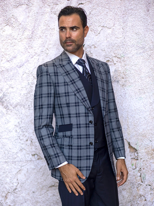 A man in the INSOMNIA PARIS-1 3PC Compose Tailored Fit Navy Suit—featuring a plaid blazer, vest, and tie—stands composedly against a textured white wall, showcasing an effortlessly tailored fit.