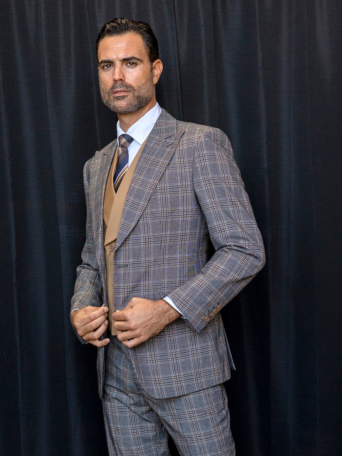 A man dressed in the INSOMNIA PARIS-2 3PC Compose Tailored Fit Brown Suit from Insomnia, featuring a beige vest and tie, stands composed against a dark backdrop, looking forward.