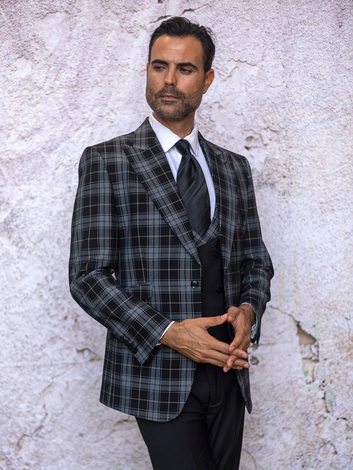 A man in an Insomnia Paris-3 3PC Compose tailored fit black suit stands against a textured, light-colored wall, hands composed and clasped.