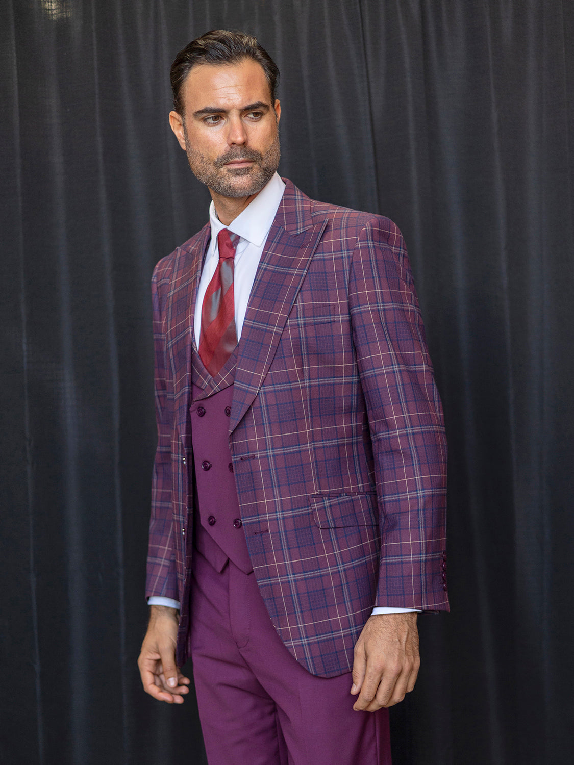 A gentleman in an INSOMNIA PARIS-3 3PC Compose Tailored Fit Burgundy Suit, from Insomnia, paired with a crisp white shirt and vibrant red tie, strikes a composed pose against a dark curtain backdrop.