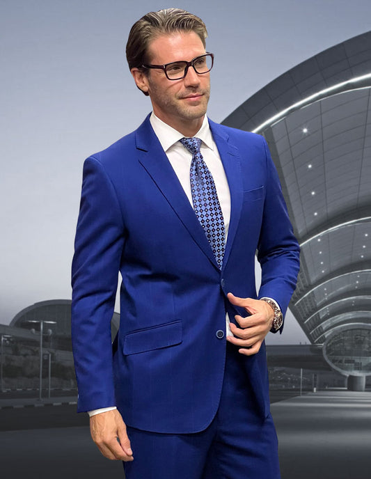 A man in a sapphire STATEMENT CLOTHING | PORTO-1-SAPPHIRE suit and glasses stands confidently against a modern architectural backdrop.