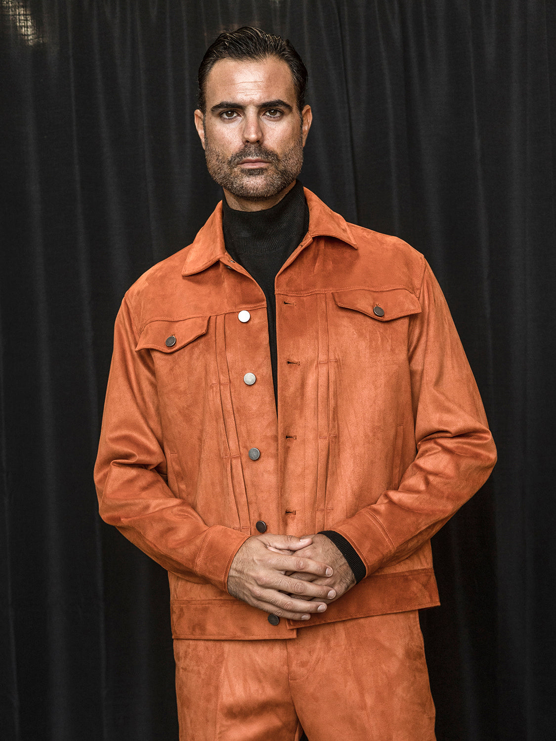 A person in an Insomnia Romeo 2PC Copper Vegan Suede Leisure Suit stands in front of a dark curtain, hands clasped.