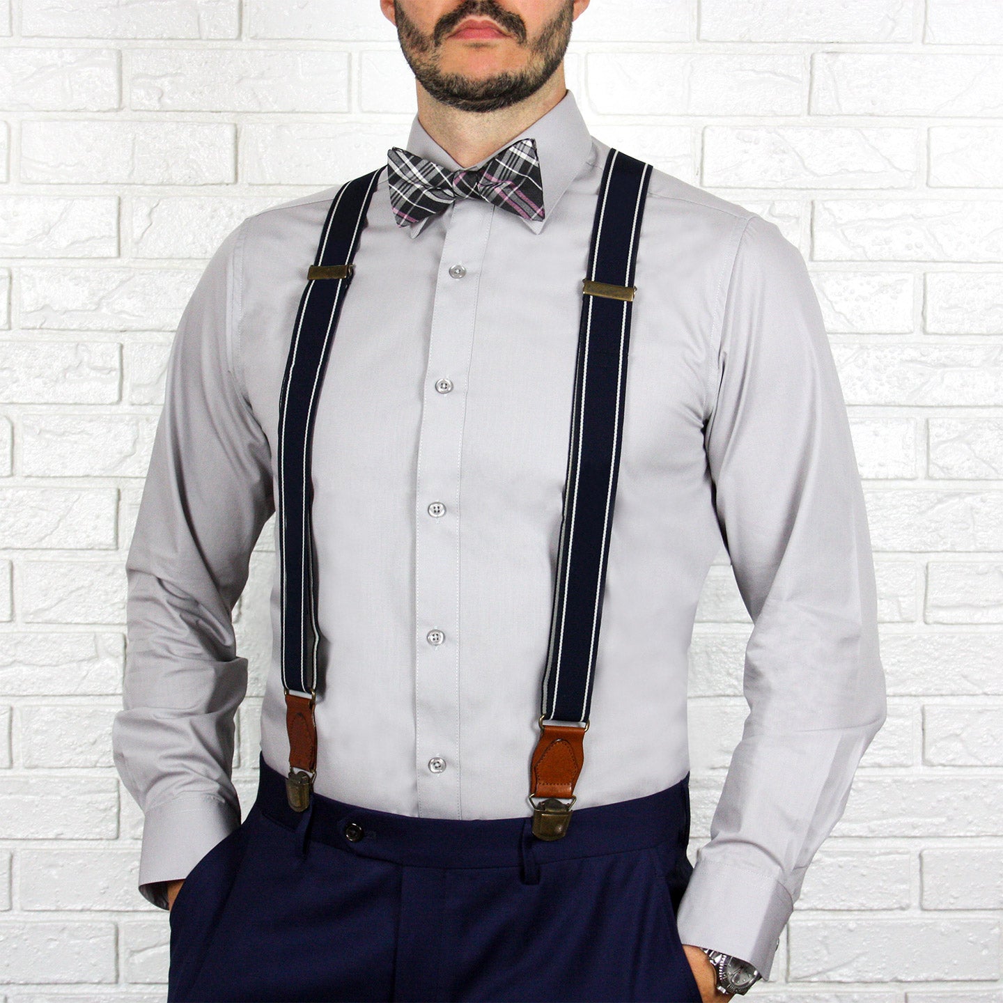 A man wearing the ARTURO Slim Fit Long Sleeve Grey Dress Shirt by Arturo, a plaid bow tie, and navy suspenders stands in front of a white brick wall.