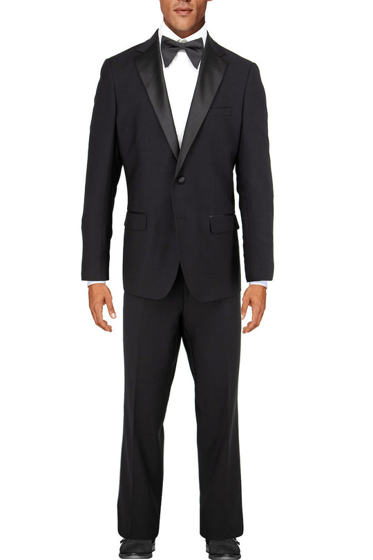 Dressed in the sophisticated Tiglio Luxe Porto Slim Fit Tuxedo Tig1001, crafted from premium Italian wool, a man stands against a plain background. The sleek European fit of his black tuxedo, paired with a classic bow tie, radiates elegance and timeless style.