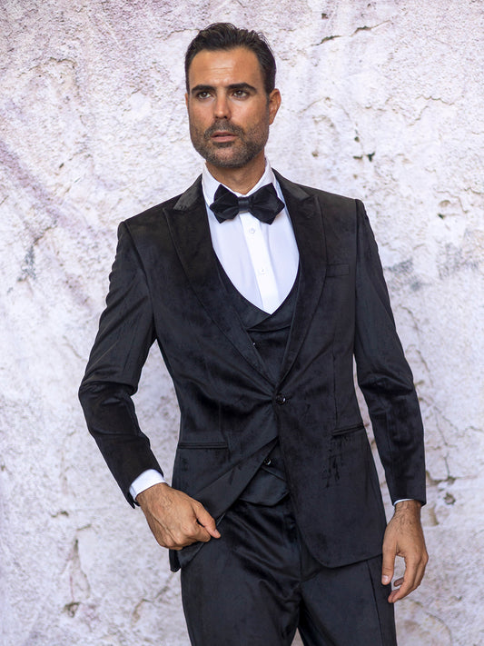 A man wearing the INSOMNIA WYNN 3PC Soft Velvet Tailored Fit Black Suit is positioned against a textured wall, gazing to the side.