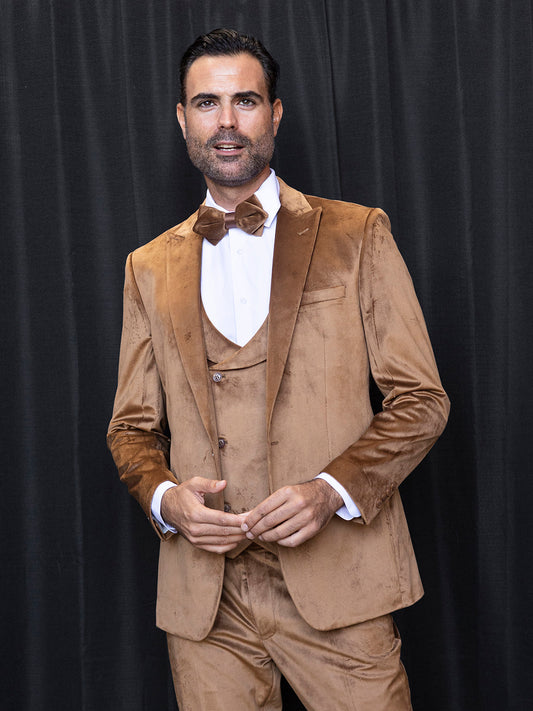 A man in an INSOMNIA WYNN 3PC Soft Velvet Tailored Fit Chestnut Suit and bowtie stands confidently in front of a black curtain, gazing directly at the camera.