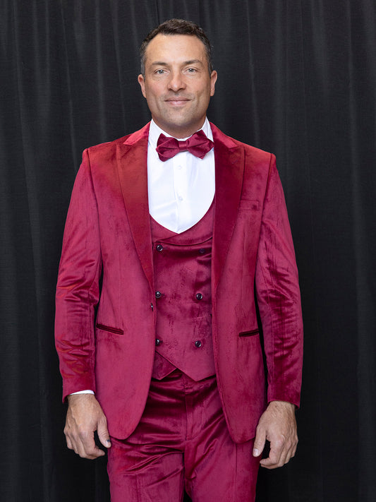 A man in an INSOMNIA WYNN 3PC Soft Velvet Tailored Fit Wine Suit stands elegantly against a black curtain, featuring a bow tie.