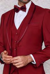 A man dressed in the INSOMNIA SUNSET 3PC Slim Fit Burgundy Tuxedo Suit by Insomnia, complete with a bow tie and reversible vest over a crisp white shirt. Confidently adjusting his suit jacket with one hand, he exudes style and poise.