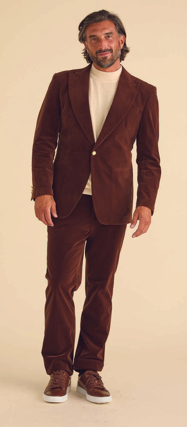 A man dressed in Inserch's Swiss Chocolate Velvet Blazer, featuring a peak lapel, paired with a white shirt and brown shoes, stands against a beige background.