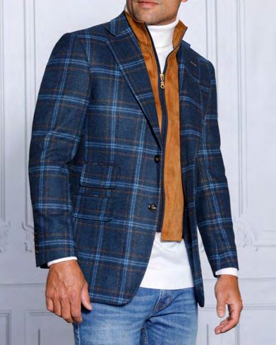 A man dressed in the Insomnia MZW-535 Tailored Fit Indigo Wool Blend Blazer, along with a white turtleneck and a brown zip-up vest featuring a removable front liner, pairs his outfit with blue jeans while standing against a light-colored paneled wall.