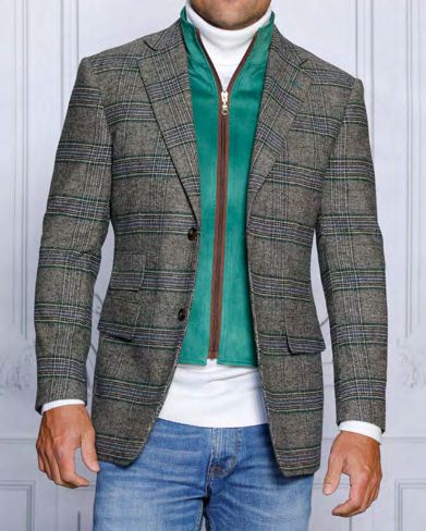 An individual displays a sharp look in the INSOMNIA MZW-535 Tailored Fit Kelly Green Wool Blend Blazer, worn over a green zip-up jacket with its removable front liner. This ensemble is perfectly complemented by a white turtleneck and blue jeans against a simple, light-colored wall.