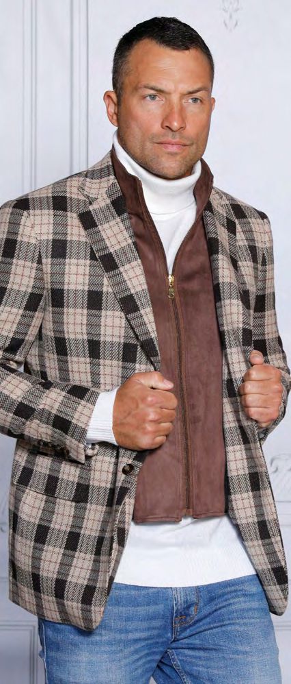 A person showcases a tailored fit, wearing the INSOMNIA MZW-536 Tailored Fit Brown Wool Blend Blazer over a plaid jacket with a removable front liner, paired with a white turtleneck sweater and blue jeans, posing against a light-colored wall.