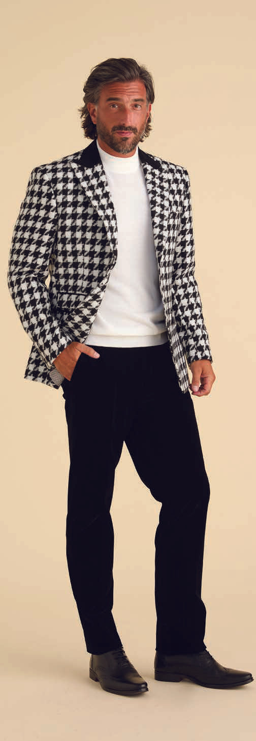 A man with gray hair and a beard is stylishly dressed in the Inserch Wool Blend Houndstooth Check Peak Lapel Blazer BL264-41 in black and white, which features modern suede trim. He elegantly pairs it with a white turtleneck sweater, black pants, and black shoes while posing against a plain background.