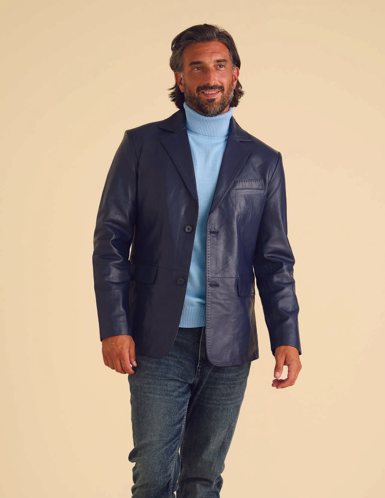 A man wearing the Inserch Lamb Leather Blazer 600-11 in navy, with its subtle pick-stitching details, pairs it stylishly with a light blue turtleneck sweater. He stands against a beige background, smiling warmly.