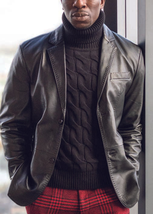 A person wearing the Inserch Lamb Leather Blazer 600-01 in black, featuring subtle pick-stitching, over a black turtleneck sweater and red plaid pants, stands indoors.