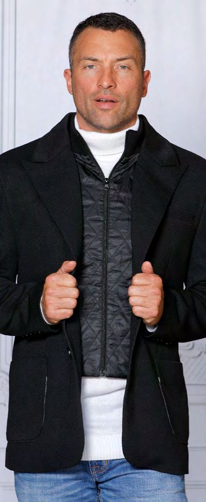 A person stands against a light background, confidently showcasing the tailored fit of the INSOMNIA SWITZERLAND Tailored Fit Black Tweed Look Blazer by Insomnia. They wear it over a patterned inner layer, paired with a white turtleneck and blue jeans, holding the blazer's lapels assertively.