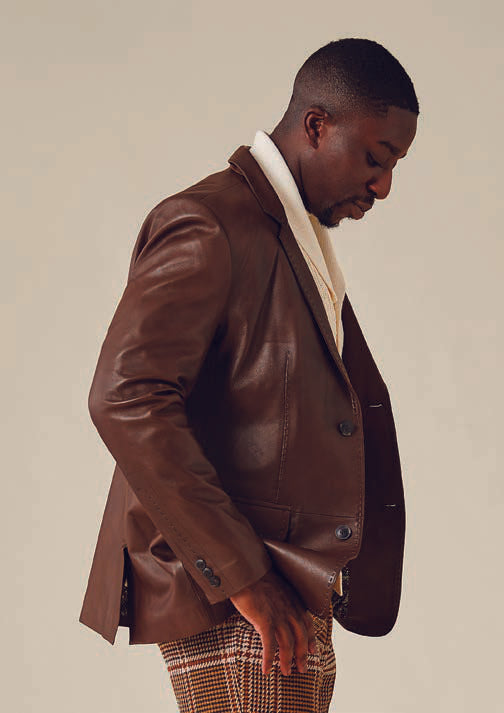 A man wearing the Inserch Lamb Leather Blazer 600-133 / 104 Cognac with a printed lining and plaid, regular-fit pants stands against a plain background, looking downward.