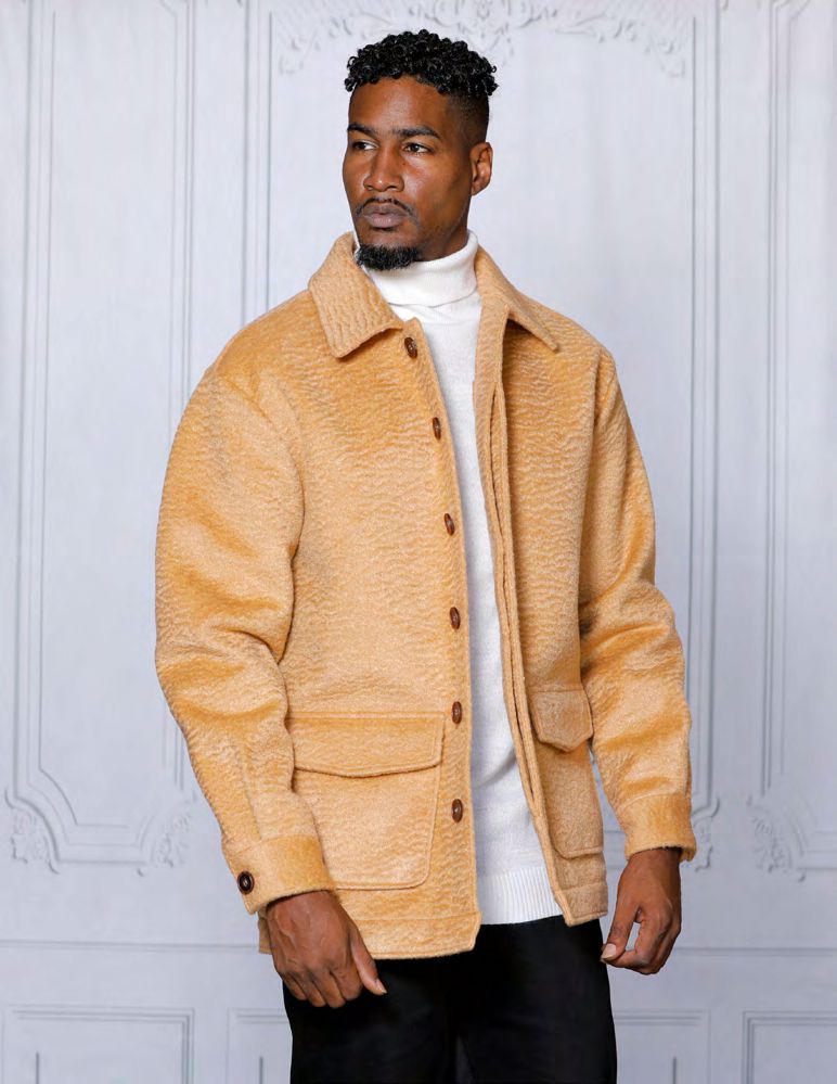 A man stands against a light patterned wall wearing the INSOMNIA CANADA Camel Poly Blend Button Coat by Insomnia, paired over a white turtleneck and black pants.