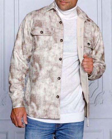 A man dressed in an INSOMNIA VALENTINO jacket in tie-dye beige tones layered over a white turtleneck sweater, paired with blue jeans featuring vegan suede accents from the Insomnia brand, stands against a white paneled wall.