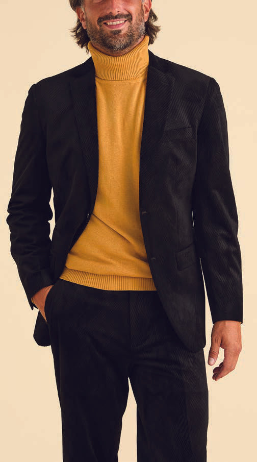 A person dressed in an Inserch Velvet Diagonal Blazer (BL275-01) in black, paired with a mustard turtleneck, stands against a beige background, smiling with hands in pockets.