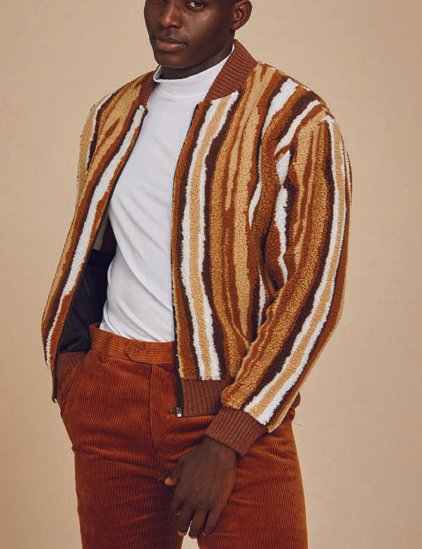 A person stands against a plain background wearing the Inserch Faux Sherpa Stripe Baseball Sweater Jacket SW610-121 Earth, featuring a striped brown and beige knit design with a baseball rib collar, layered over a white shirt and paired with brown pants.