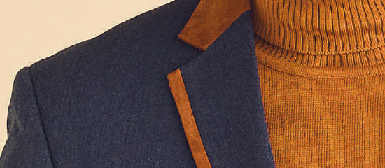 A man dressed in the Inserch Denim Blazer BL274-10 in denim blue, complemented by brown accents and a brown turtleneck, stands against a neutral backdrop, exuding both elegance and style.