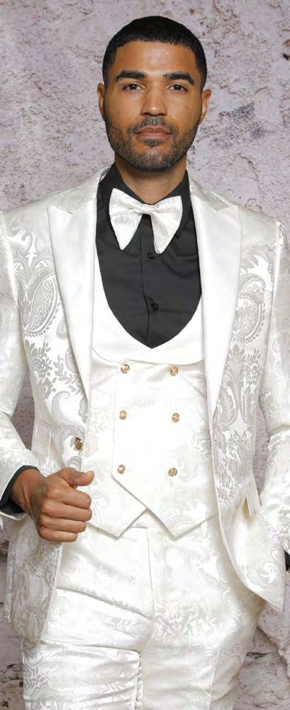 A man wearing an INSOMNIA ARMANI Slim Fit Cream Fancy Woven Suit from the Insomnia brand, paired with a white vest and a black shirt featuring a large bow tie, stands against a textured wall.