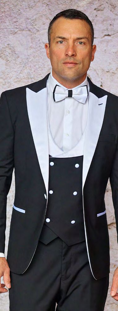 Dressed in the sleek Insomnia ROME 3PC Slim Fit Black White Spandex Tuxedo Suit and a matching bow tie, a man confidently stands against a textured wall, gazing directly at the camera.