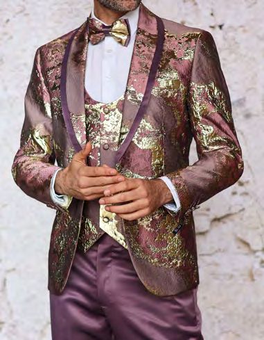 A person dressed in the INSOMNIA DELMAS 3PC Slim Fit Maroon Fancy Woven Suit by Insomnia, featuring an intricate gold and purple pattern, complemented with a matching vest and bow tie, posed against a light background.