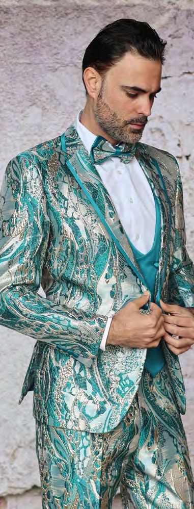 A man in the INSOMNIA SANTO 3PC Tailored Fit Turquoise Suit by Insomnia, featuring teal and gold patterns, along with a matching bow tie and vest, adjusts his jacket while standing against a textured wall.