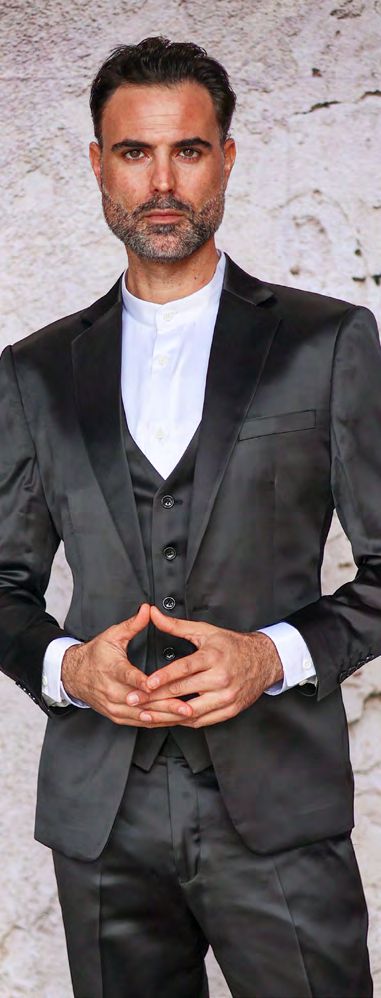 A man in an INSOMNIA CLASSICO 3PC Tailored Fit Satin Spandex Black Suit and a white shirt stands in front of a textured, light-colored wall. He has short dark hair and a beard, with his hands lightly clasped.