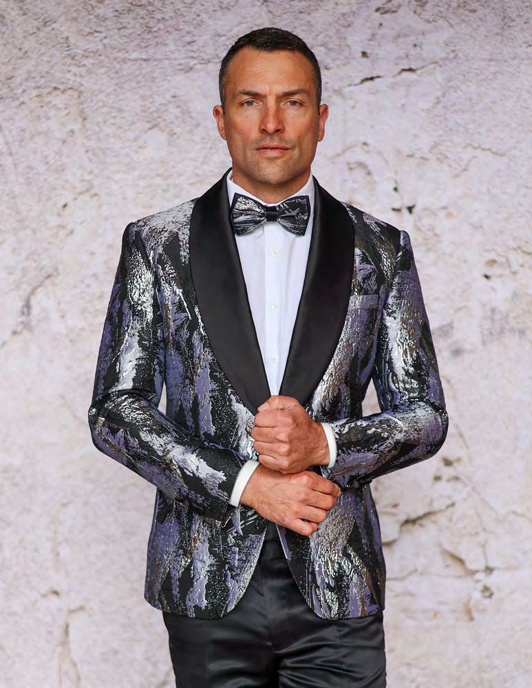 A man wearing an Insomnia INSOMNIA MZS-552 Slim Fit Silver Woven Sport Coat with a black lapel and bow tie stands against a textured wall background.