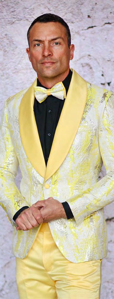 Standing against a light-colored stone background, the person exudes elegance in an Insomnia slim-fit yellow and silver patterned suit with a matching bow tie and black shirt, completed by the sleek INSOMNIA MZS-552 Slim Fit Yellow Woven Sport Coat.