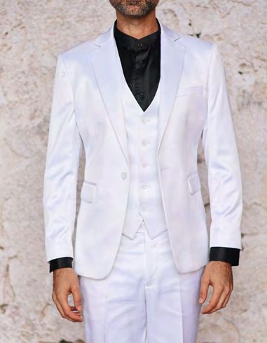 A man, elegantly attired in the INSOMNIA CLASSICO 3PC Tailored Fit Satin Spandex White Suit from Insomnia, featuring a black shirt and tie, stands against a textured, light-colored wall. The subtle sheen of satin spandex adds an extra touch of sophistication to his ensemble.