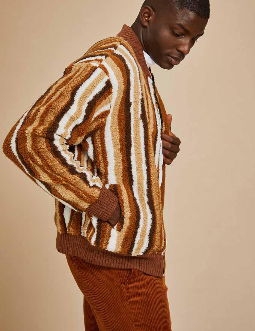 A person stands against a plain background wearing the Inserch Faux Sherpa Stripe Baseball Sweater Jacket SW610-121 Earth, featuring a striped brown and beige knit design with a baseball rib collar, layered over a white shirt and paired with brown pants.