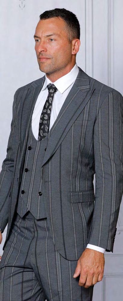 A man dressed in the INSOMNIA ITALIA 3PC tailored fit charcoal suit by Insomnia, paired with a white shirt and patterned tie, stands against a light-colored wall.