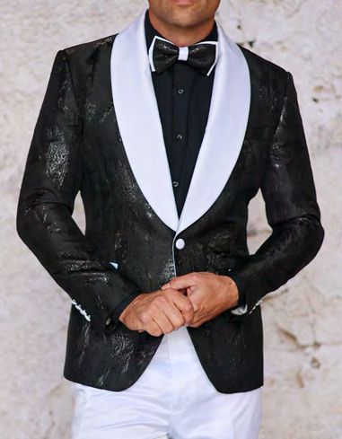 A person wearing the INSOMNIA MZS-552 Slim Fit Black Woven Sport Coat with a shiny texture, distinct white lapels, and a black bow tie stands against a textured stone background, hands elegantly clasped in front.