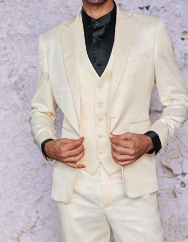 An individual is wearing the INSOMNIA CLASSICO 3PC Tailored Fit Satin Spandex Cream Suit from Insomnia, complete with a coordinating satin vest and a black shirt, standing against a textured background.