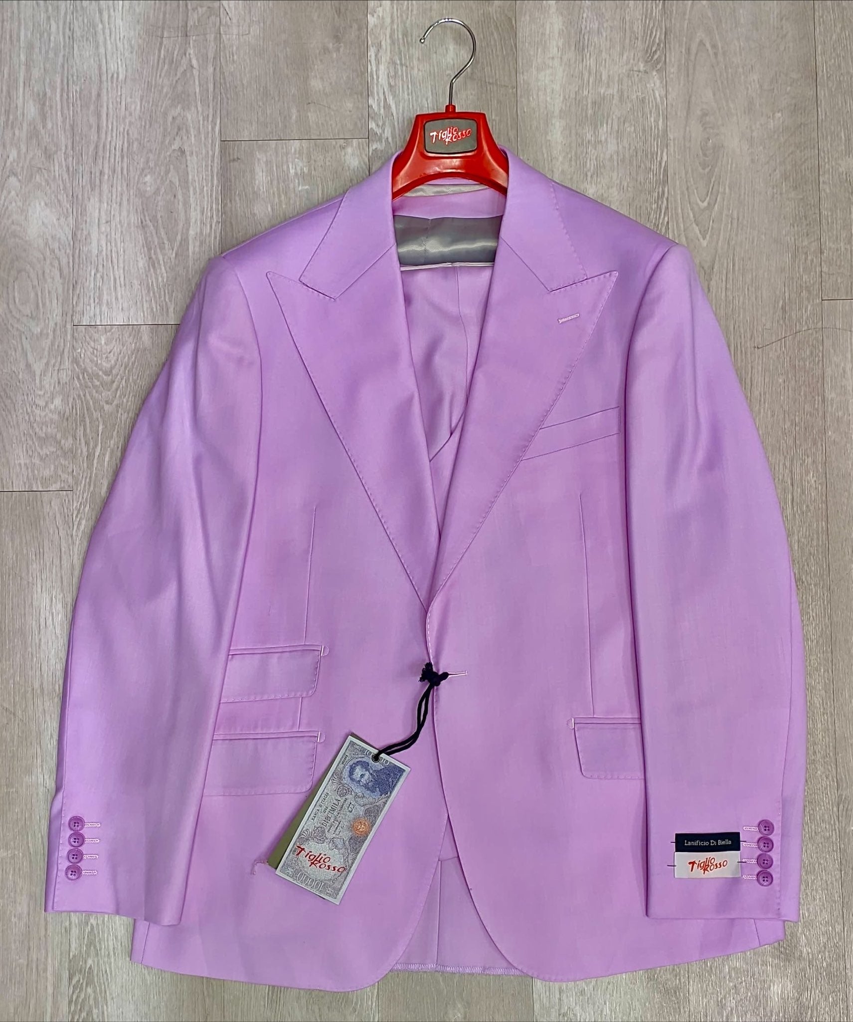 A Tiglio Rosso Orvietto solid lilac wool suit jacket with notched lapels, part of a sophisticated three-piece ensemble (size 54L only), rests elegantly on a wooden floor. Its attached tag ensures authenticity and quality, highlighting its luxurious 100% wool fabric.