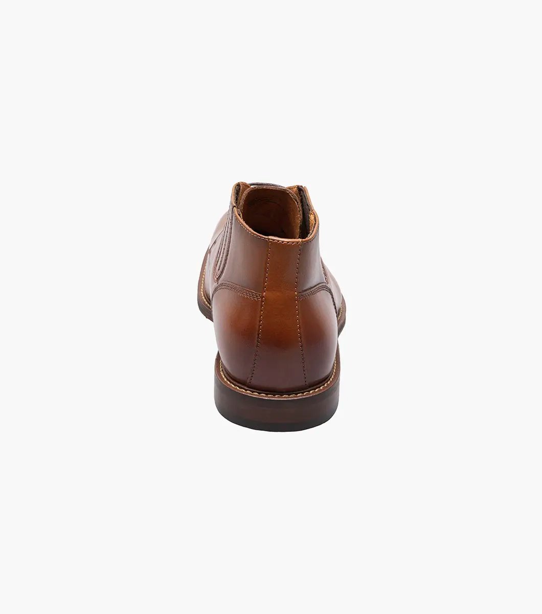 The Stacy Adams Maxwell Plain Toe Chukka Boot in Cognac features a sophisticated leather design with three eyelets, detailed stitching, and a versatile low heel, making it ideal for both dressy and casual events.