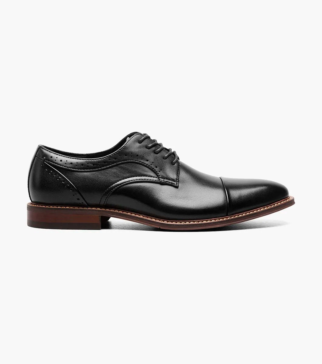 The Stacy Adams - MADDOX Cap Toe Oxford shoe features a black smooth leather finish with a polished appearance, complemented by a brown sole, intricate decorative stitching, and anatomical arch support.