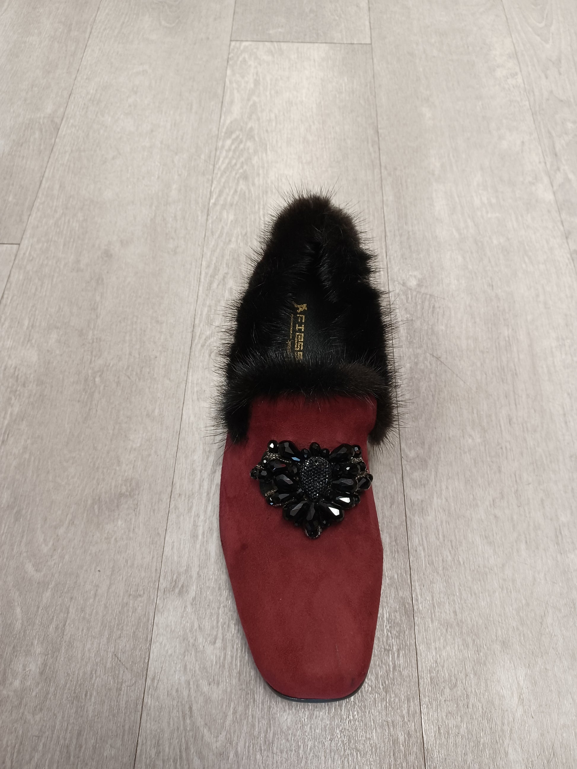 The Fiesso Shoe #202, a red velvet slipper adorned with black fur trim and a black beaded heart embellishment, rests elegantly on a light wood floor. Please note: this item is part of our final sale collection and is available only in sizes 8 and 12 with no returns.