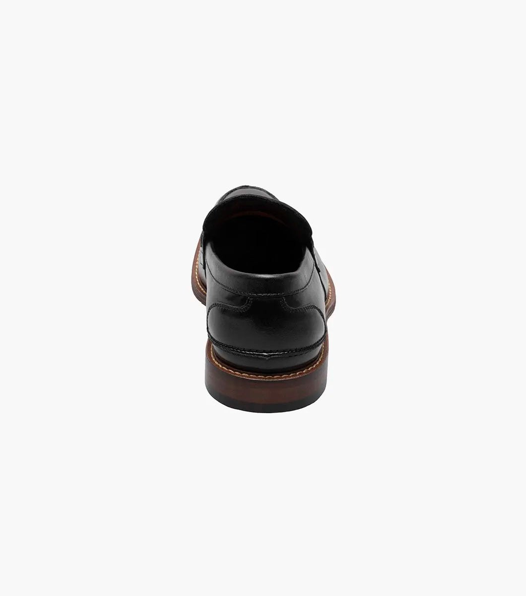 The Stacy Adams MARLOWE Algonquin Moc Toe Penny Slip On in black, featuring a refined leather upper and a contrasting brown sole, is displayed against a plain background.