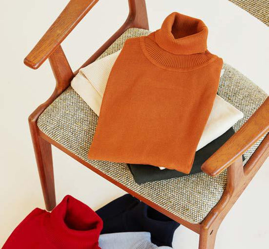 Inserch's cotton blend turtleneck sweaters, part of the 4708 collection featuring ten color options, are elegantly displayed. The vibrant orange, white, and green variations rest on a wooden chair with a patterned cushion, while other regular fit options in red, blue, and black are arranged gracefully on the ground below.